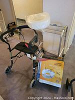 Nexus black metal walker with padded seat and backrest, plastic-wrapped seat back, and red strap, next to foldable metal walker frame and Guardian elevated toilet seat box.