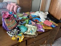 Collection of assorted fabric scarves in multiple colors and patterns spread on a wooden surface with three purses including a purple purse with multicolor geometric front and tan handle, a black purse, and a light beige purse.