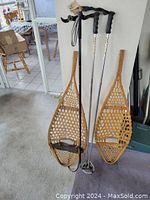 Pair of vintage wooden snowshoes and two metal snow poles shown standing upright against a wall on carpeted floor.