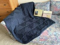 Photo showing black knit throw blanket draped over floral patterned sofa with two packaged sheet sets placed on the sofa; one Ralph Lauren in beige and gold packaging.