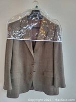 Front view of a brown wool men's blazer on hanger covered partly by plastic cover. Shows tailored fit, notch lapel, two-button closure, and front flap pockets.