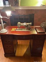 Front view of sewing machine desk with lamp, showing cream-green Necchi sewing machine embedded in wood desk with seven drawers and dark wood finish.