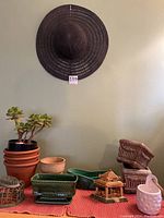 Wide view showing black woven garden hat hung on wall, jade plant on table, terra cotta pots stacked, green planters, floral frogs, Chinese pagoda, and other small garden items.