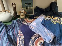 Overview of lot including Japanese apron, linen garment, two tablecloths, ceramic items including Noritake teapot, clay vase and cat mug, arranged on a surface with a framed print in background.