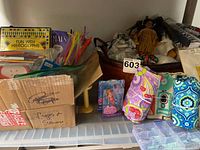 Wide shot showing a box with 'Cups & Spoons', colorful pipe cleaners in a clear bowl, a Native American style doll, and rolled items including aprons and a tablecloth.