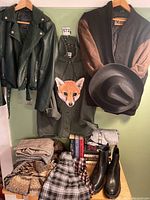 Clothing items hanging and arranged on shelf, including jackets, a hat, folded plaid pants and skirts, books, and black ankle boots.