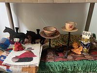 Photo showing various rustic items including black metal horse candlesticks, Pisa plates, tea cups, a small carved wooden angel, butterfly vase, black swan figurine, and textile items such as pillow covers and table runner.