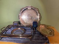 Full view of the four silverplate items on a blue tablecloth including the round tray, rectangular casserole holder, diamond-shaped dish, and round glass bowl with silver rim alongside cleaning supplies.