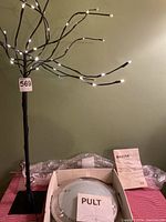 Photo of bendable light tree with multiple lit LED pin lights on black branches, standing on a table, also showing two IKEA ceiling light fixtures in packaging with installation manuals
