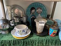 First photo showing multiple items including trays, coffee pots, ceramic vase, plates, bowl, tins on green cloth.