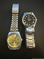 Two men's silver tone watches placed side by side on black background showing full watches and bracelets