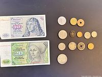 Two German Deutsche Mark banknotes (10 and 20) alongside twelve assorted foreign coins laid out on a dark surface.