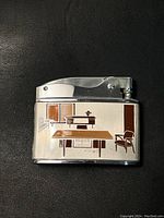 Front view of lighter showing mid-century office furniture illustration in brown and beige on chrome body.