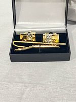 Set of gold tone cufflinks and matching tie clip in black gift box with cushioned gray interior, shown from front angle.