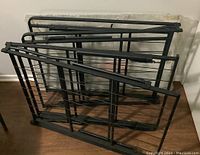 Two black metal bed frames folded and leaning against a wall, showing tubular metal side rails and wire slats for mattress support.