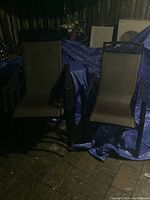 Two black metal framed outdoor chairs with brown mesh seating and backrest, placed on brick surface over a blue tarp.