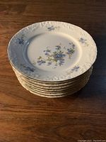 Stack of 8 white ceramic plates with blue floral pattern and embossed edges under warm lighting.