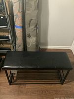 Side angled view of the black bench showing the cushioned top and metal slat lower shelf. Shows wear on the top surface and overall construction.