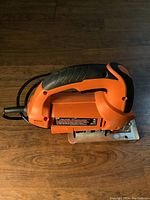 Side view of Black & Decker jigsaw showing orange body with black grip and power cord; metal base plate visible beneath jigsaw blade area.