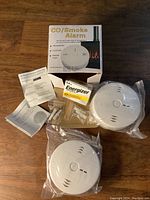 Lot contents shown including two sealed CO/Smoke alarms, product box, instruction manuals, mounting hardware, and Energizer AA batteries.