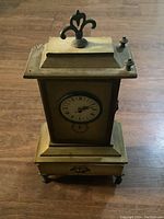 Front and top view of brass-finished battery operated clock jewelry box showing clock face and decorative finials on top.