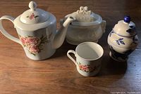 Full set of fine china tea accessories arranged on a wood surface: teapot with pink roses, matching teacup, white sugar bowl with lid, and blue and white ginger jar.