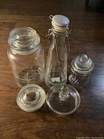 Five glass canisters and jars arranged on floor showing various lid types and bottle shapes