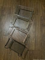 Four metal wire baskets stacked inside each other on a wooden floor. Different finishes including dark, bronze-like and black.
