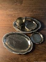 Photo displaying all silver items on wooden floor including two oblong trays, two round coasters, and a footed cup