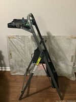 Side angled view of the painters ladder showing the metal frame, black textured steps, and black top tool tray with compartments.