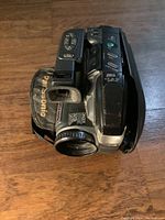 Top angled view of the vintage Panasonic handheld video camera showing control buttons, lens, hand strap with Panasonic branding, and part of the carrying pouch.
