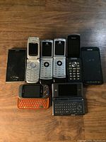 Photo showing all eight phones laid out on a wooden surface, displaying variety of models and condition, including cracked screen and keypad styles.