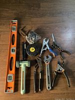 Photo showing an orange spirit level, a green mallet, large pipe wrench, abrasive wire wheel brushes, pliers, adjustable wrenches, scissors, and hand pruners arranged on a wooden floor.