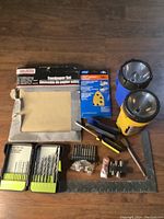 Various sanding and hardware items including a sandpaper set, drill bit set, flashlights, screwdriver, hardware screws, protractor ruler and utility knife laid out on wood surface.