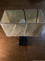 Top-front angle view of lamp showing rectangular beige shade with plastic cover, light bulb inside, and black base.