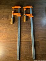 Two steel bar clamps laid side by side on wooden surface showing full length with orange clamp arms and wooden handles.