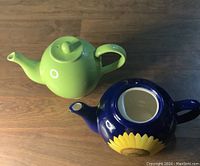 Photo showing green ceramic teapot and blue ceramic teapot with sunflower on wooden surface.