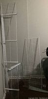 Photo showing three white metal wire shelving units with metal brackets leaning against a wall.