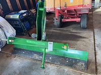Front and side view of 5ft green steel swing scraper blade attachment with 3-point hitch bracket and support stand on workshop floor.