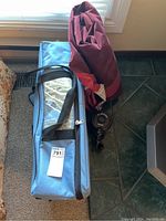 Portable bed frame in blue carrying case alongside rolled burgundy Hillary air mattress with attached black air pump.