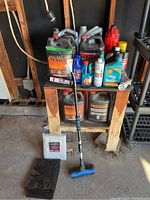 View of assorted vehicle oils, coolants, car care sprays, and a snow brush arranged on and below a wooden table in a garage setting.