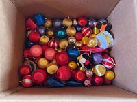 Box filled with assorted Christmas tree bulbs in multiple colors and shapes including round, teardrop, striped, and solid colors along with a yellow plush ornament with a face design.