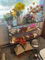 Wide view of the decorative lot showing multiple arrangements of artificial flowers in assorted vases and containers, the matching table lamps, and the decorative milk can style waste bin.