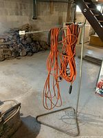 Metal rod stand holding multiple orange extension cords in a basement/workshop environment with concrete floor and wood stacks nearby.