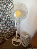 Full view of Airworks floor fan standing next to wall with floral wallpaper and power outlet visible, showing its base, pole, and fan head.