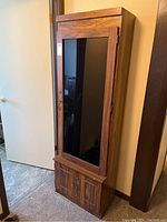 Front angled view of the tall wooden firearm cabinet with tinted glass door and lower storage compartment with two doors.