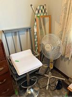 White vinyl bar stool, metal shoe rack, ironing board with patterned cover, standing fan, wall mirror, and iron all arranged in a corner with curtain backdrop.
