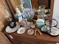 Overview of assorted decor items on a wooden dresser including a black floral hurricane lamp, decorative plates, lighthouse tealight holder, small glass jars, and other decor pieces.