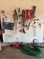 Wide shot showing red hydraulic car jack on the ground, coiled green garden hoses, several hand tools hanging on white pegboard including jumper cables, orange extension cords, bungee cords, pruning shears, loppers, and manual tools.