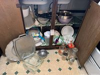 Overview of kitchen items inside a wooden cabinet and on the floor including trays, glass dishes, plastic containers, strainers and glass jars